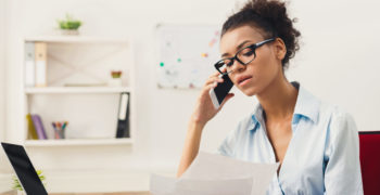 Young serious african-american businesswoman talking by phone with papers, sitting at modern office workplace. Business consulting, copy space